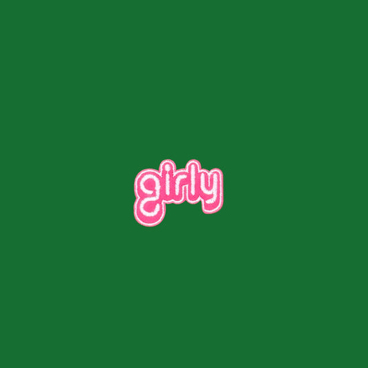 Girly written in lowercase script in white on a pink outline border embroidered iron-on patch on a solid green surface. 